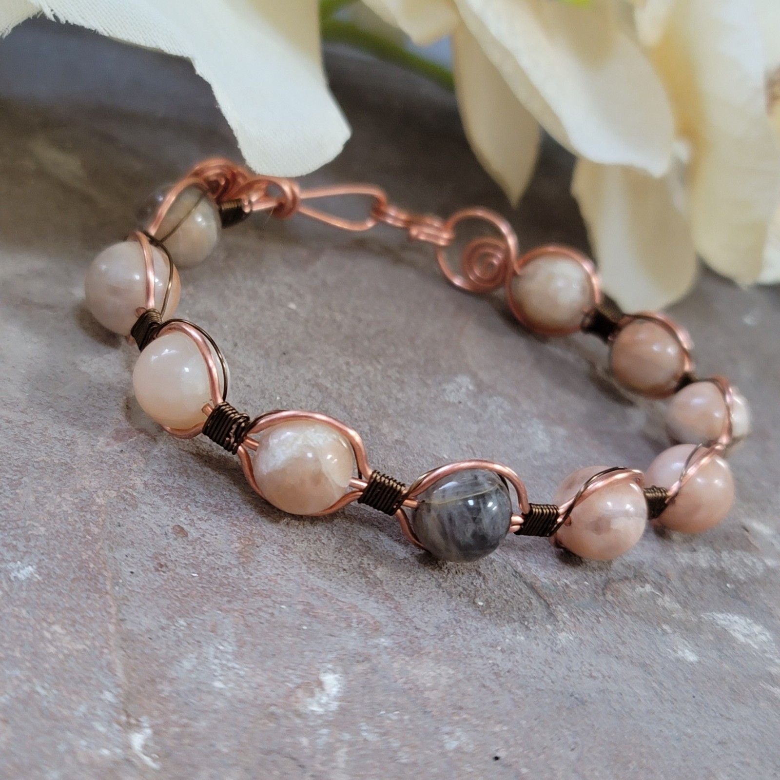 Bracelet with pink beads and copper wire on a stone surface with white flowers in the background