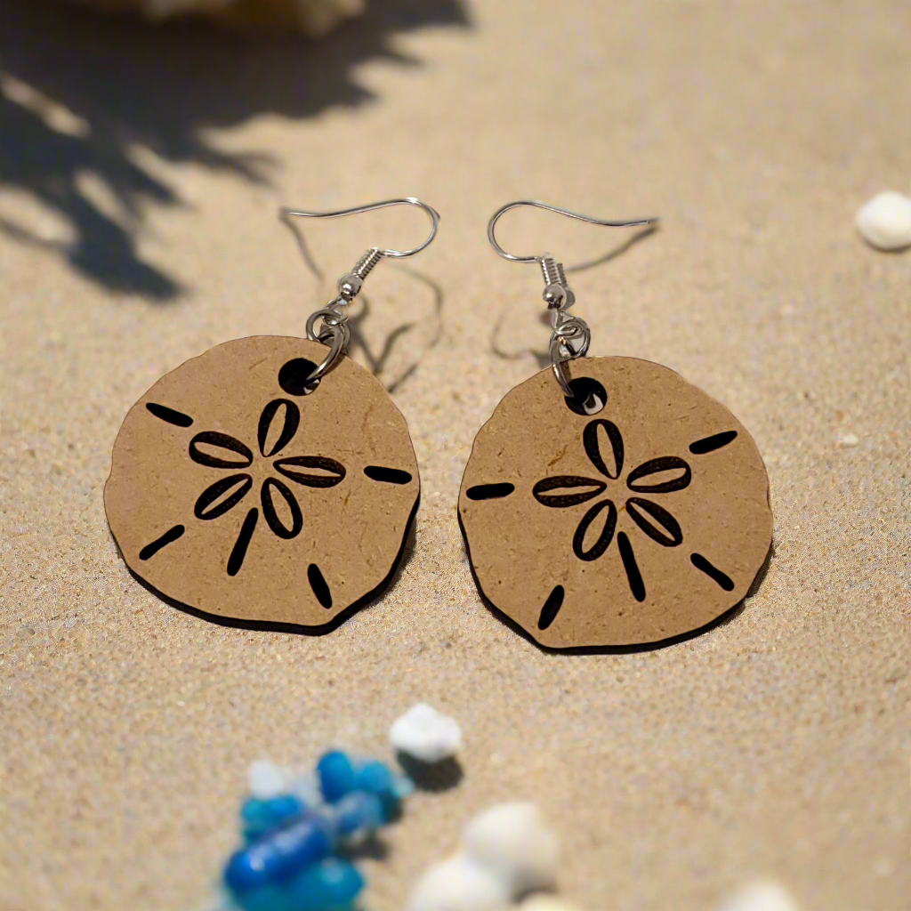 A pair of handmade laser cut sand dollar dangle earrings laid on a sandy surface with seashells and small stones around.