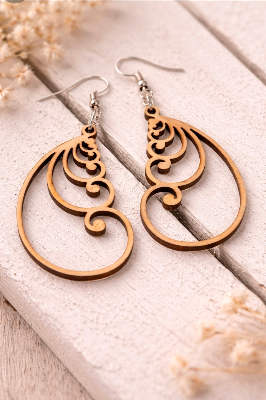 Close-up photo showing a woman wearing a lightweight teardrop-shaped wooden statement earring with swirling cutout design. The earring hangs from a silver hook and rests just above the jawline. The background is softly blurred to highlight the jewelry.