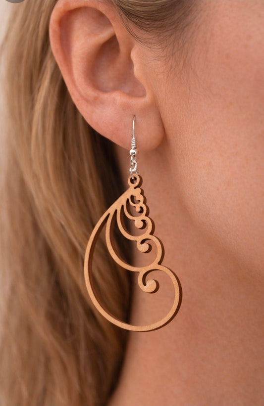 Close-up photo showing a woman wearing a lightweight teardrop-shaped wooden statement earring with swirling cutout design. The earring hangs from a silver hook and rests just above the jawline. The background is softly blurred to highlight the jewelry.