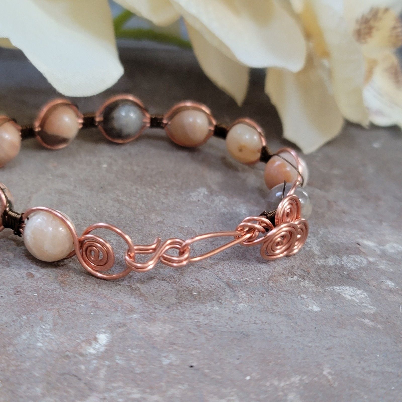 Bracelet with beads and copper wire design on a stone surface with flowers in the background