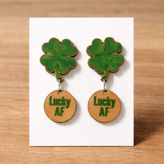 Pair of green four-leaf clover earrings with 'Lucky AF' on a white card against a wooden background